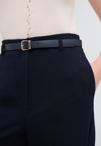 Černý kožený pásek se zlatou sponou, nošený přes tmavě modré kalhoty s hladkou texturou. Obsahuje ribbený, světle béžový top s knoflíky.