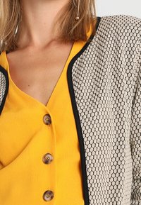 Veste à motif noir et crème avec une finition texturée, associée à un haut jaune avec un col en V et des détails à boutons.