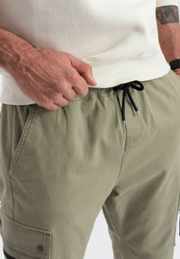 Męska ręka poprawia pas biodrowy oliwkowych szortów cargo z czarnym sznurkiem, na sobie ma kremowy sweter i czarny zegarek na nadgarstku.