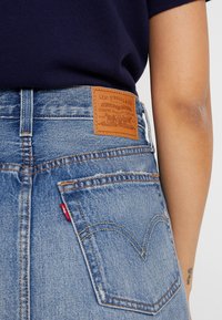 Blue denim jeans with a brown leather patch featuring brand details. Distressed texture and classic pocket design are visible.