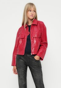 Giacca cropped in pelle sintetica rossa con colletto eDue tasche sul petto, indossata sopra una camicia nera e abbinata a pantaloni neri a trama.