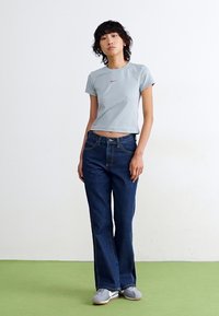 Lyseblå cropped T-shirt med rødt Nike-logo, parret med mørkeblå flared jeans. Modellen har grå sneakers på og står på et grønt gulv.
