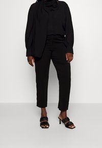 Svart blus med knäppning, lös passform och scarfdetalj, i kombination med skräddarsydda svarta byxor. Svarta klacksandaler med volangdetaljer.