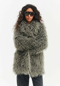 Kobieta z długimi kręconymi włosami, nosząca duże, ciemne okulary przeciwsłoneczne oraz gruby, szary, kudłaty płaszcz ze skrzyżowanymi ramionami.