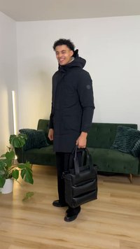 Juoda pūkinė striukė su kapišonu, turi du priekinės kišenes ir logotipo ženklelį. Laiko juodą struktūruotą rankinę. Vidaus patalpos su žalia sofa.