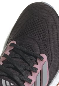 Svart atletisk sko med en texturerad stickad överdel, rosa detaljer på snörningssystemet och en vit sula med mönstrad profil.