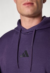 Sweatshirt roxa com capuz ajustável, apresentando um pequeno logotipo preto da Adidas no peito e um tecido macio e texturizado.