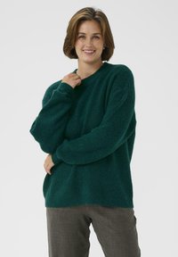 Grüner, strukturierter Strickpullover mit langen Ärmeln und rundem Ausschnitt. Über grauen Hosen getragen, die eine lockere, entspannte Passform haben.