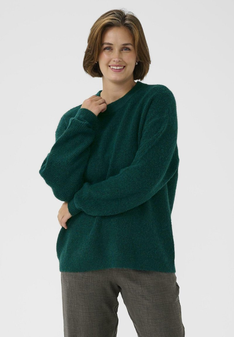 Grüner, strukturierter Strickpullover mit langen Ärmeln und rundem Ausschnitt. Über grauen Hosen getragen, die eine lockere, entspannte Passform haben.