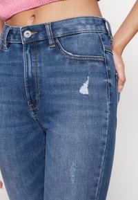 Primer plano de una persona que lleva puestos unos vaqueros de tiro medio de color azul con detalles desgastados y un top corto de punto rosa, con una mano en el bolsillo.