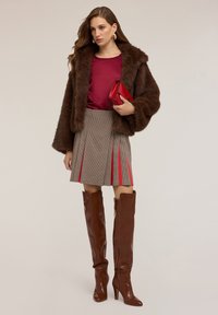 Giacca in finta pelliccia marrone, blusa bordeaux, gonna a pieghe a quadretti con accenti rossi e stivali marroni con tacco al ginocchio. La borsa a tracolla rossa completa l'outfit.