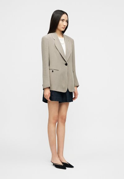 Γυναίκα που στέκεται, φορώντας ένα μπεζ ριγέ σακάκι με ένα κουμπί, λευκή μπλούζα, σκούρα σορτς και μαύρα μυτερά παπούτσια slingback, μπροστά σε απλό φόντο.