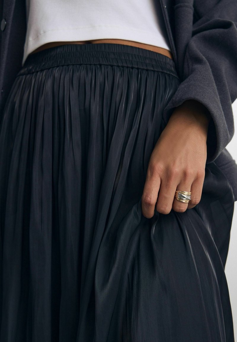 Main portant une bague en or à plusieurs anneaux tenant un tissu de jupe plissée noire, montrant la ceinture ainsi qu'une partie d'un haut blanc et d'une manche de veste sombre.