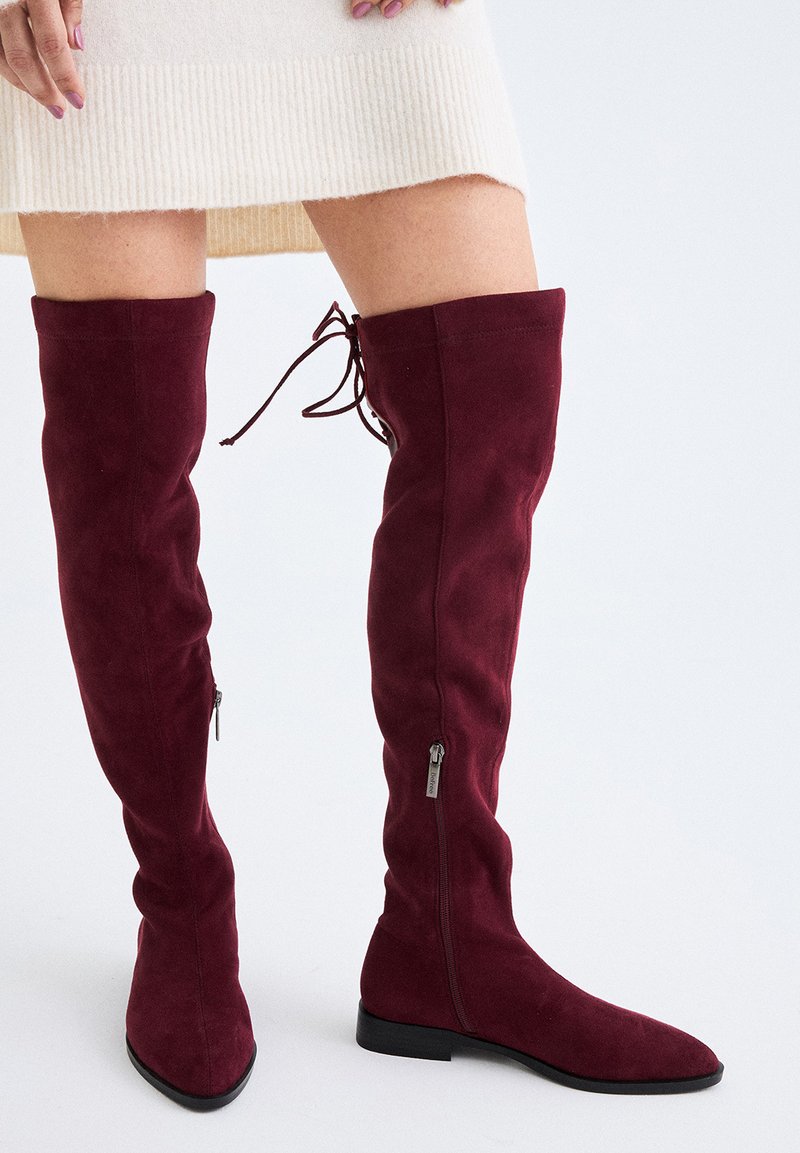 Mujer llevando botas de ante burdeos hasta la rodilla con cremalleras laterales y lazos en la parte trasera, combinadas con una falda de punto acanalado crema.