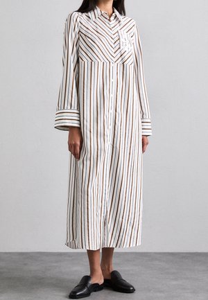 Kvinde iført en hvid langærmet kjole med knapper ned foran, med lodrette brune og sorte striber, og sorte slip-on sko, stående på en grå gulv.