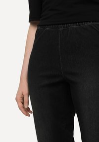 Pantalones de denim negro con una banda elástica en la cintura, textura sutil y detalles de costura en contraste en las costuras. Una mano se apoya ligeramente sobre la tela.