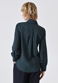 Dunkelbluse in Teal mit Kragen, langen Ärmeln und Knopfmanschetten. Verfügt über eine Rückenfalte und eine glatte Textur, gestaltet für eine lockere Passform.