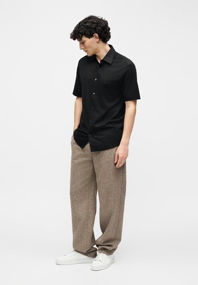 Άντρας που φοράει μαύρο πουκάμισο με κοντά μανίκια και κουμπιά, χαλαρά καφέ παντελόνια και λευκά αθλητικά παπούτσια, στέκεται με τα χέρια στις τσέπες κοιτώντας προς τα κάτω.