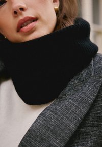 Crni pleteni džemper s ovratnikom s rebrastom teksturom, slojevit ispod sivog teksturiranog blejzera i svijetle majice. Zlati detalji su vidljivi.