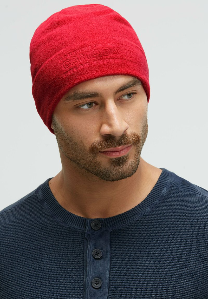 Red fleece beanie with textured surface and embossed logo. Man wears a navy blue textured henley shirt with three buttons.