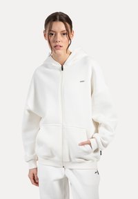 Hvid zip-up hoodie lavet af blød stof, med en forlommer, ribbede mansjetter og en hætte. Enkel design med en lille logo accenter.