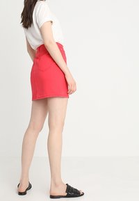 Jupe mini en denim rouge avec une taille haute, deux poches arrière et un ourlet effiloché. Assortie avec un t-shirt blanc et des sandales noires à enfiler.