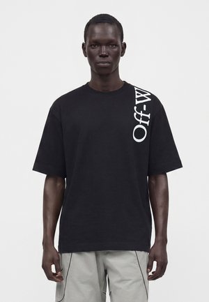 Homme portant un t-shirt noir oversize avec un texte blanc vertical "Off-White" sur l'épaule droite et un pantalon gris clair, debout devant un fond uni.
