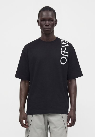 Άνδρας που φοράει μαύρο oversized μπλουζάκι με κατακόρυφο λευκό κείμενο "Off-White" στον δεξί ώμο και ανοιχτό γκρι παντελόνι, στέκεται μπροστά σε απλό φόντο.