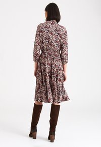 Robe fleurie en tissu sombre avec des motifs roses, bleus et blancs. Présente des manches trois-quarts, une taille élastique et un ourlet froncé. Bottes marron jusqu'aux genoux.