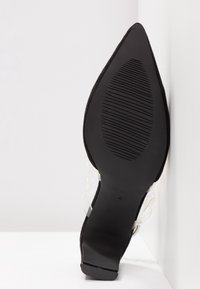 Chaussure à talon haut en daim noir avec un bout pointu et des brides transparentes. La semelle noire texturée présente des rainures pour une meilleure adhérence.