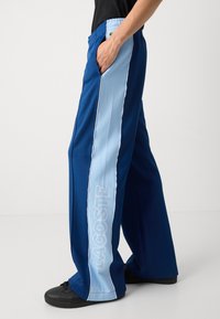 Pantalones azules con franjas laterales en azul claro, adornados con ribetes blancos y "LACOSTE" en letras grandes. Tela suave, corte relajado y bolsillos laterales.