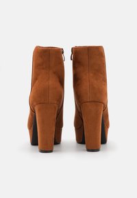 Bottines à talons en suede marron avec une texture lisse et une fermeture éclair à l'arrière, présentant un talon large et un design à bout rond.