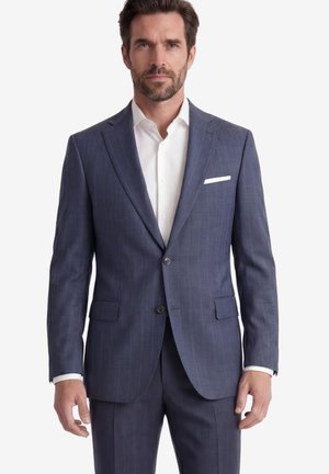 Mann steht frontal und trägt eine blaue karierte Anzugjacke, passende Hose und ein weißes Hemd.