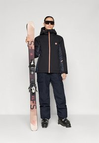 Veste de ski noire avec fermeture éclair à l'avant et accents rose clair, associée à un pantalon de ski cargo foncé et des chaussures de ski noires. Tenant des skis roses avec fixations.