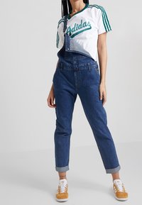 Henkilö, jolla on päällään valkoinen Adidas-t-paita vihreillä raidoilla, siniset farkkuhaalarit, joista yksi olkonauha on alas, sekä sinapinkeltaiset Adidas-tenniskenkä.