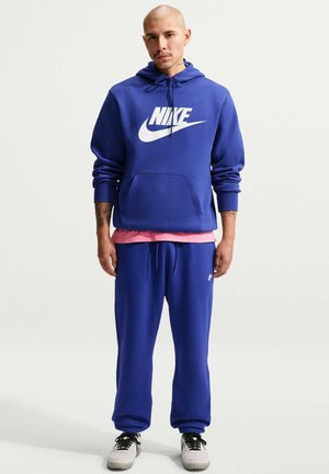 Man draagt een blauwe Nike hoodie en matching joggingbroek, roze shirt eronder en witte sneakers met zwarte veters, staand tegen een witte achtergrond.