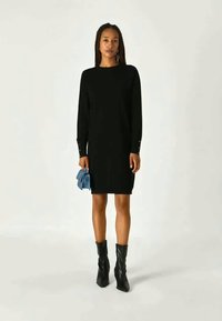 Vestido de malha preto com mangas longas, punhos canelados com botões decorativos. Modelo segura uma clutch azul clara, combinada com botas altas pretas.