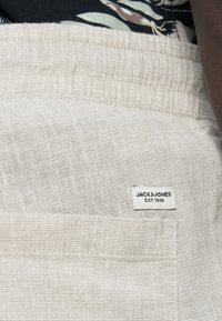 Ανοιχτό μπεζ ύφασμα με υφή υφάσματος, που περιλαμβάνει μια ετικέτα με την επιγραφή "JACK & JONES EST.1990." Σχέδιο με χαλαρή εφαρμογή και ελαστική μέση.
