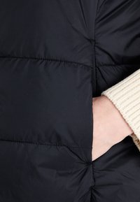 Main dans une manche de pull côtelé beige insérée dans la poche latérale d'une doudoune matelassée noire.