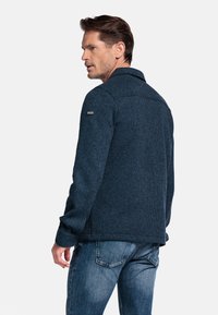Navyblauwe textuurjack met kraag en lange mouwen, met een merkaanduiding op de linkerschouder. Draag het met jeans.
