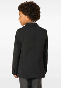 Kiharatuksega poiss, kes kannab musta bleiserit ja halli pükse, nähtud tagant, suunaga vasakule, lihtsa valge tausta ees.