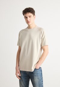 Beige tričko s raglánovými rukávy vyrobené z měkké tkaniny, s kulatým výstřihem a decentním logem na hrudi, spárované s modrými džíny.