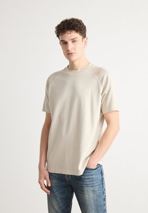 T-shirt beige à manches raglan en tissu doux, avec un col ras du cou et un logo discret sur la poitrine, associé à un jean bleu.