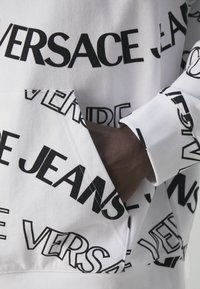 Un sweat à capuche blanc portant un motif répété de logos noirs avec "Versace" et "Jeans". Comprend une poche kangourou et des poignets côtelés.