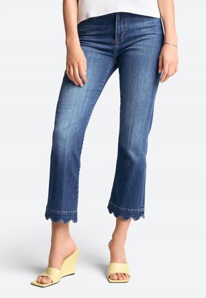 Dunkelblaue, verkürzte Denim-Jeans mit gewelltem Saum, kombiniert mit gelben Keilsandalen. Der Stoff hat eine glatte Oberfläche und eine leichte Ausbleichung.