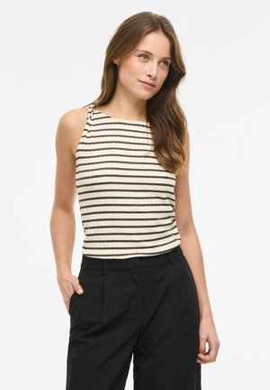 Femme portant un haut blanc sans manches avec des rayures horizontales noires et un pantalon noir taille haute, debout avec une main dans la poche.