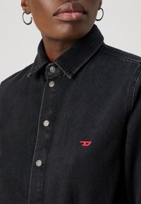 Zwart denim overhemd met een kraag, drukknoopsluiting en een rood logo op de borst. Het materiaal lijkt stevig met een soepele textuur.