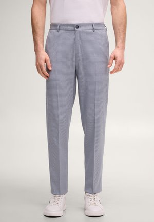 Mann trägt hellblaue maßgeschneiderte Hose und weiße Sneakers, steht vor einem schlichten hellen Hintergrund.