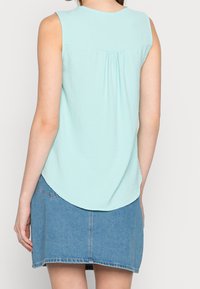 Top senza maniche di un leggero verde mente con scollatura arrotondata e pieghe posteriori, abbinato a una minigonna di denim con orlo sfrangiato.