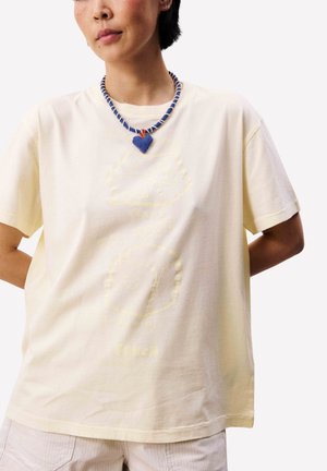 Personne portant un T-shirt jaune pâle orné de graphiques de fromage discrets et un collier en perles bleues avec un pendentif en forme de cœur.